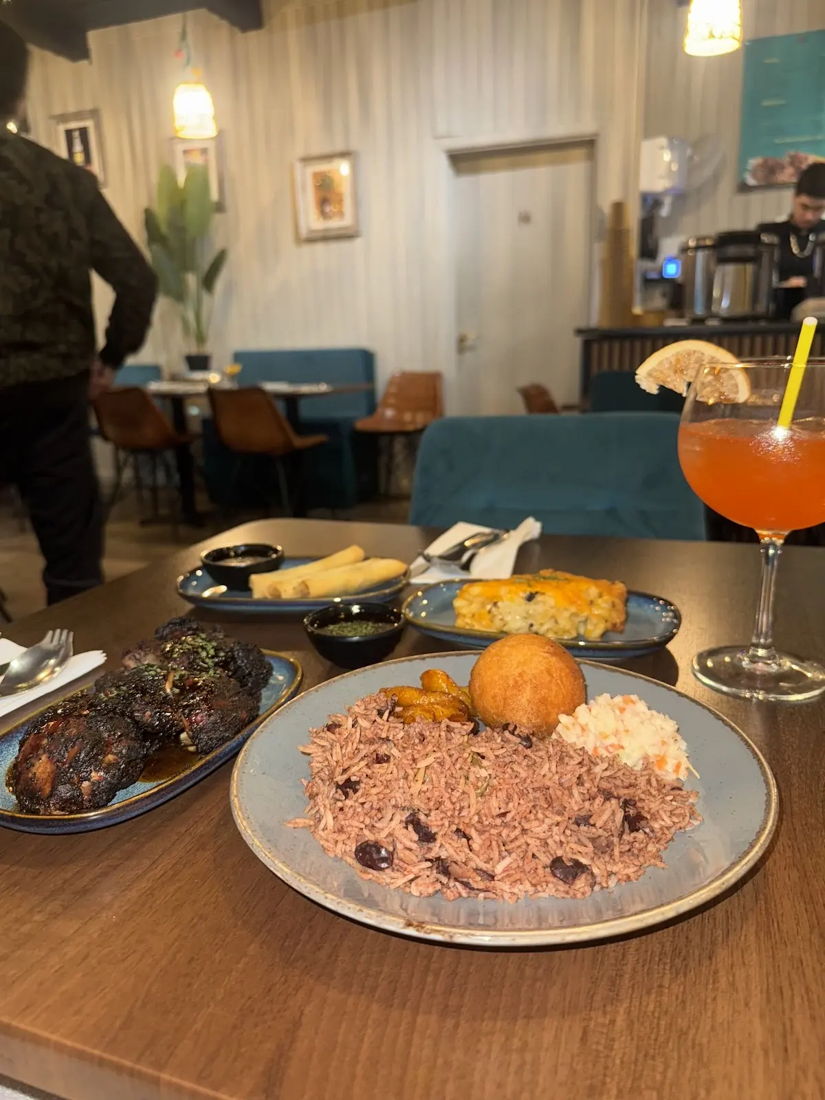 The dining room at Chel's Fusion seen past a laid table of jerk chicken, rice and peas and a cocktail, pendant lights overhead